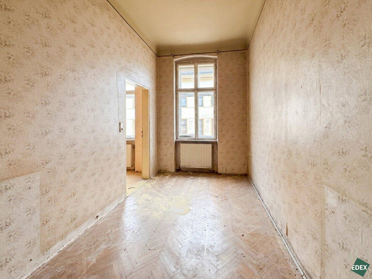 Leerer Raum mit abgenutzter Tapete, Fischgrätparkett und einem großen Fenster.
