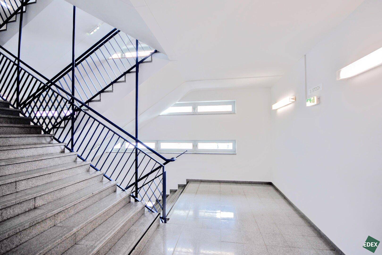 Geräumiges Treppenhaus mit Steinstufen, Metallgeländer und großen Fenstern für viel Licht.