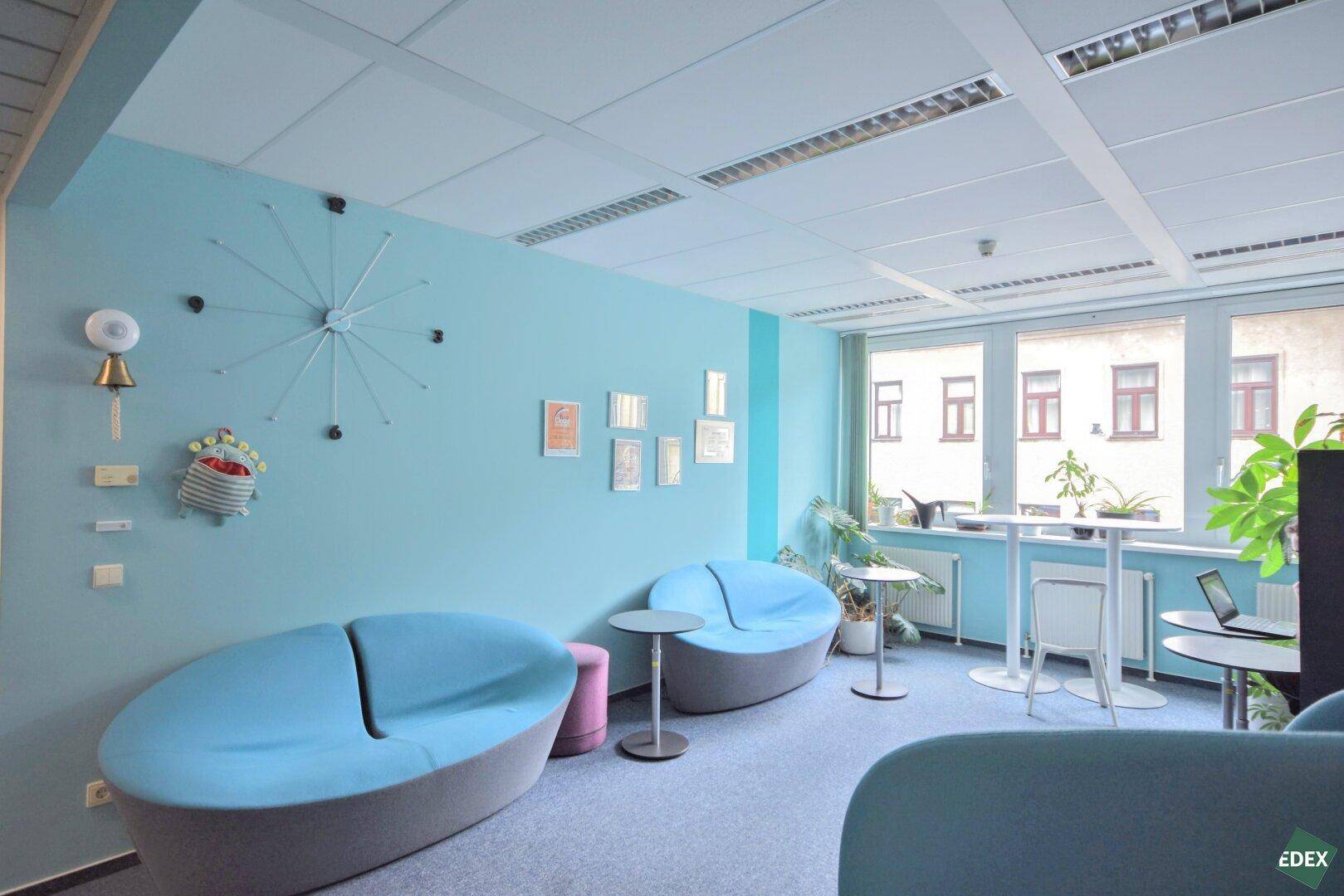 Heller Loungebereich mit blauen Sofas, kleinen Tischen und einer auffälligen Wanduhr in modernem Design.