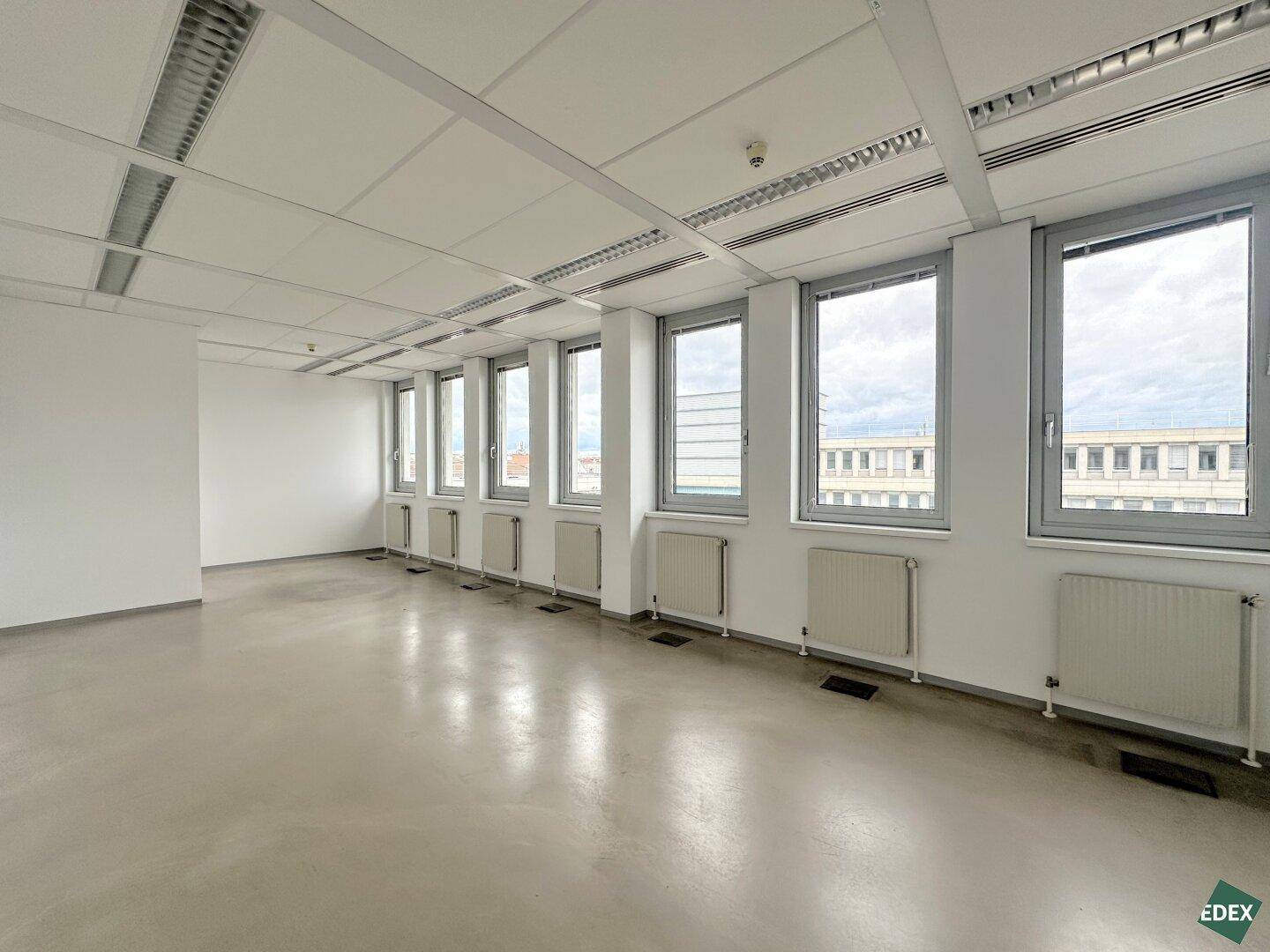 Großer, leerer Büroraum mit einer langen Fensterfront, die viel Tageslicht hereinlässt.