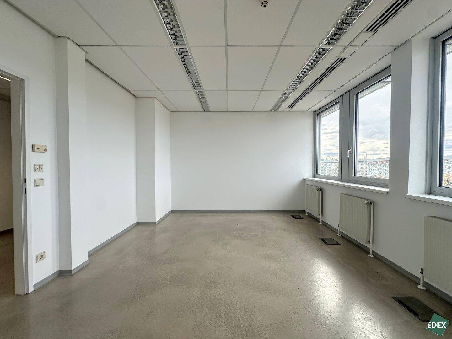 Heller Büroraum mit großen Fenstern und einem Zugang zu weiteren Räumlichkeiten.