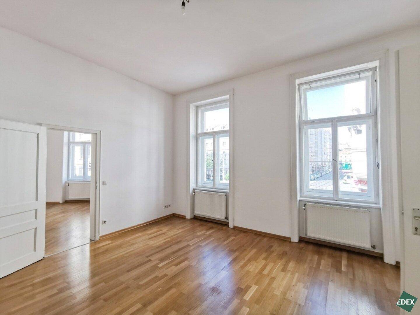 Geräumiger Raum mit Holzboden, hohen Decken und großen Fenstern mit Blick auf die Straße.