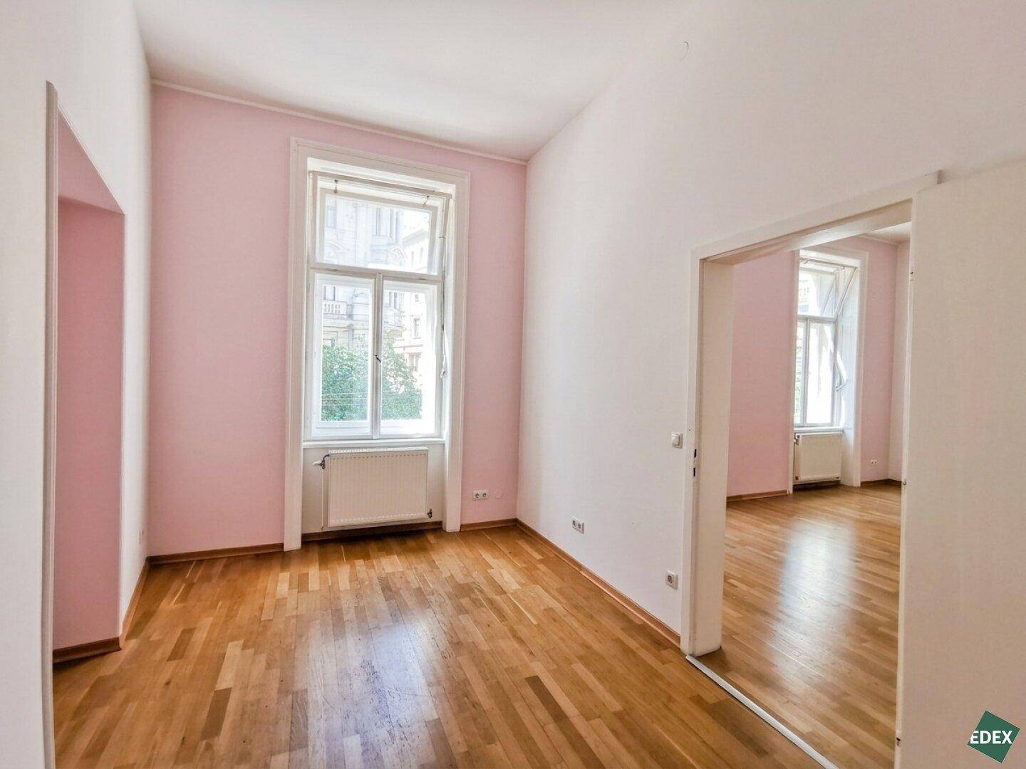 Zimmer mit rosa Wänden, Holzboden und einem großen Fenster, das den Raum erhellt.