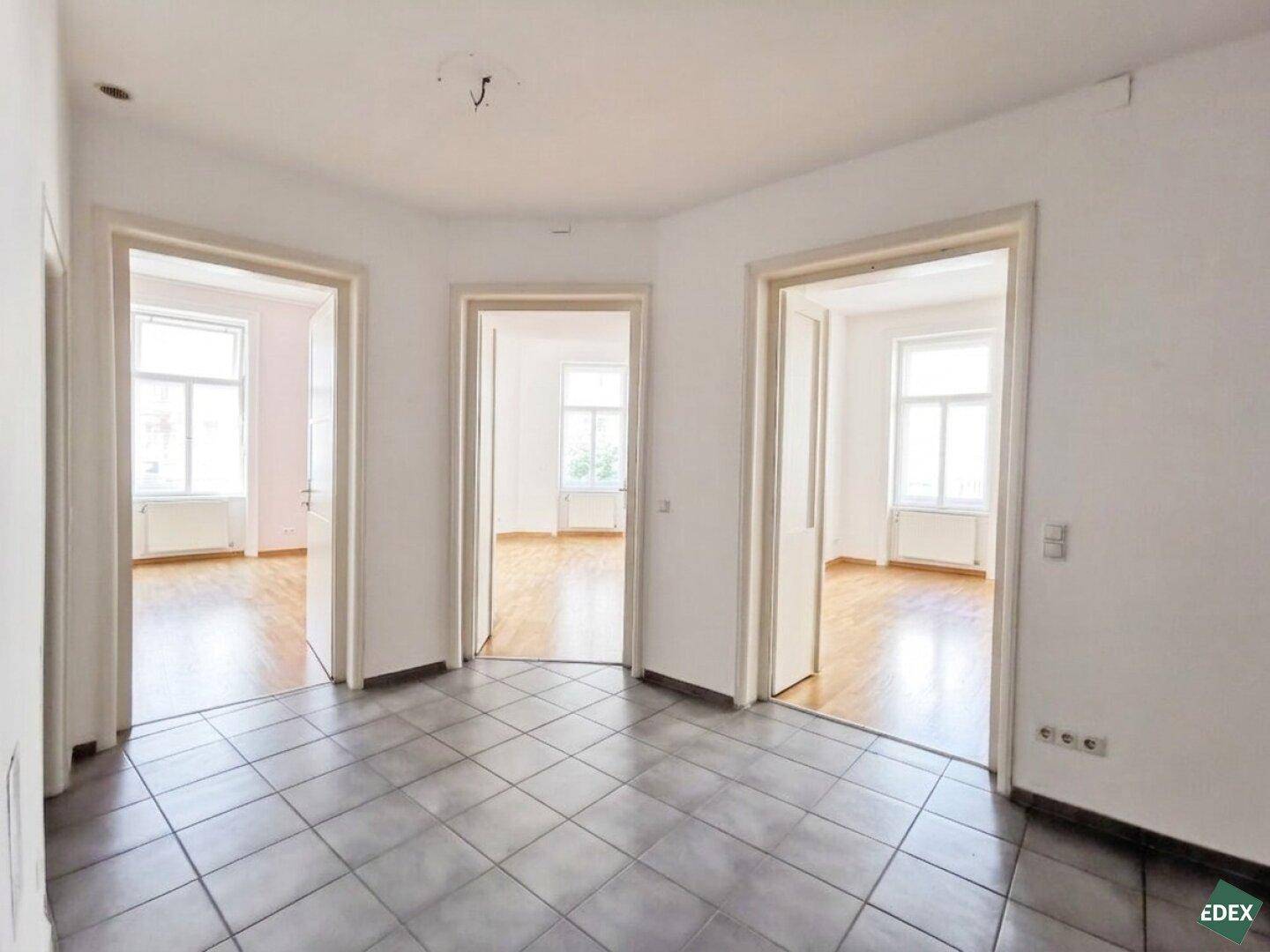 Zentraler Flur mit grauen Fliesen und drei Türen, die zu hellen Zimmern führen.