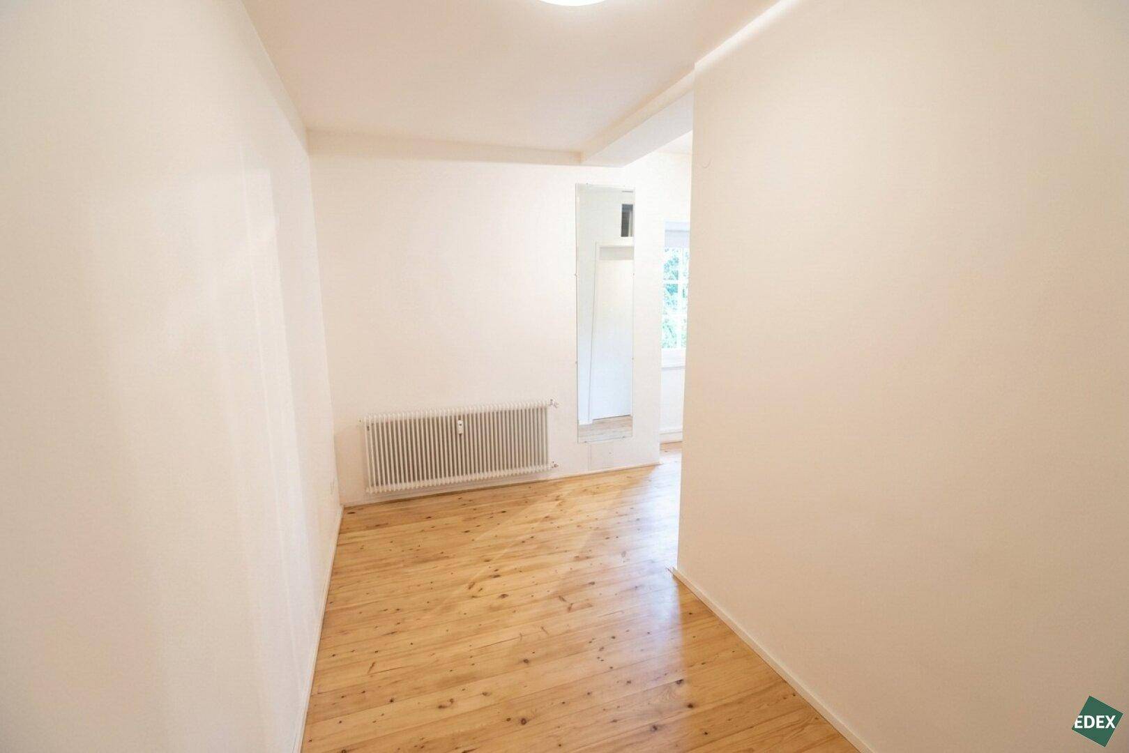 Schmaler Flur mit Holzdielenboden und weiß gestrichenen Wänden, der zu einem weiteren Bereich führt.
