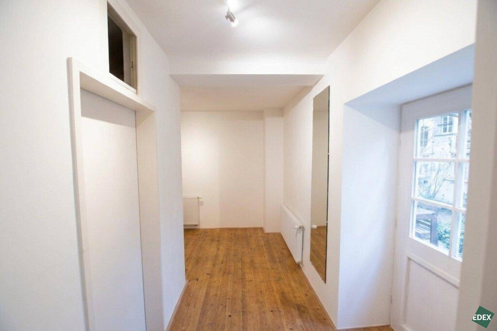 Langer, schmaler Flur mit Holzdielenboden, weiß gestrichenen Wänden und einem Fenster am Ende.