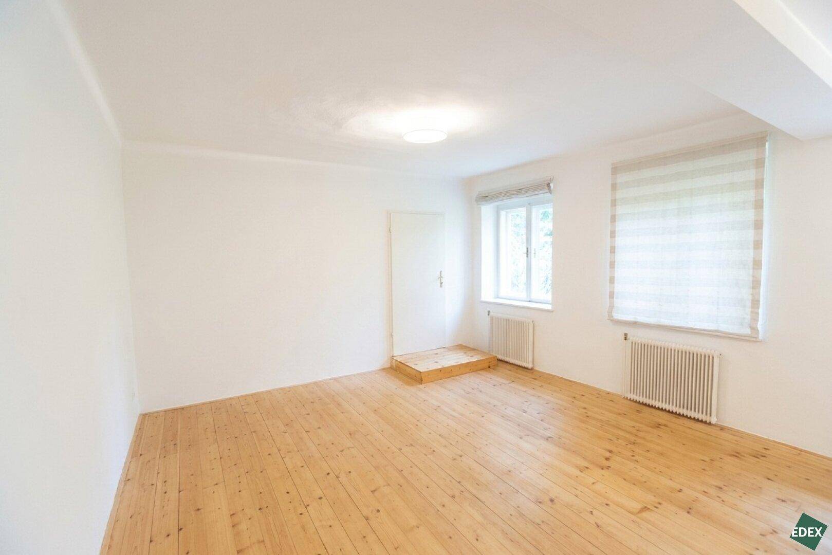 Leerer Raum mit Holzdielenboden, einem Fenster mit Jalousie und einer Tür, die zu einem erhöhten Bereich führt.