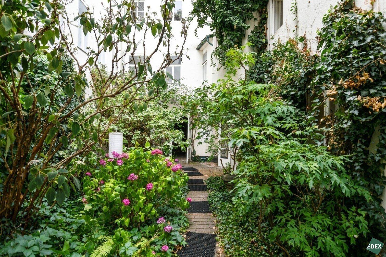Grüner Innenhof mit üppiger Vegetation, Blumen und einem schmalen Weg zwischen den Gebäuden.