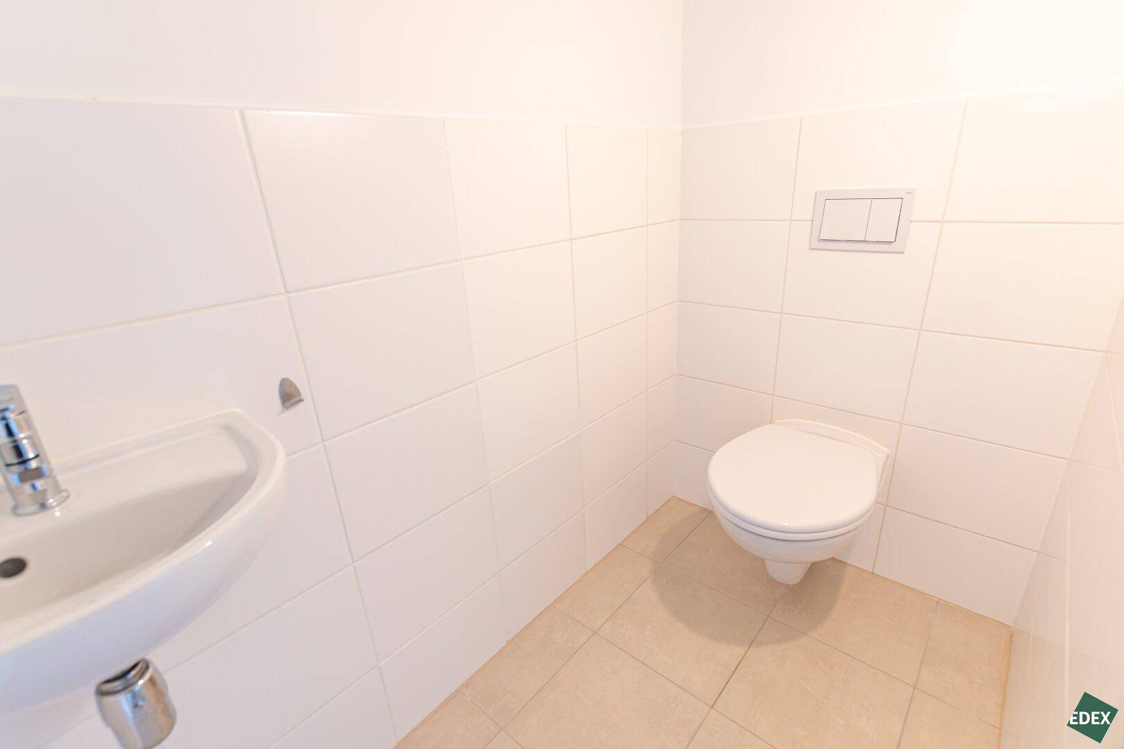 Separates WC mit wandhängender Toilette und kleinem Waschbecken, komplett gefliest in Weiß.