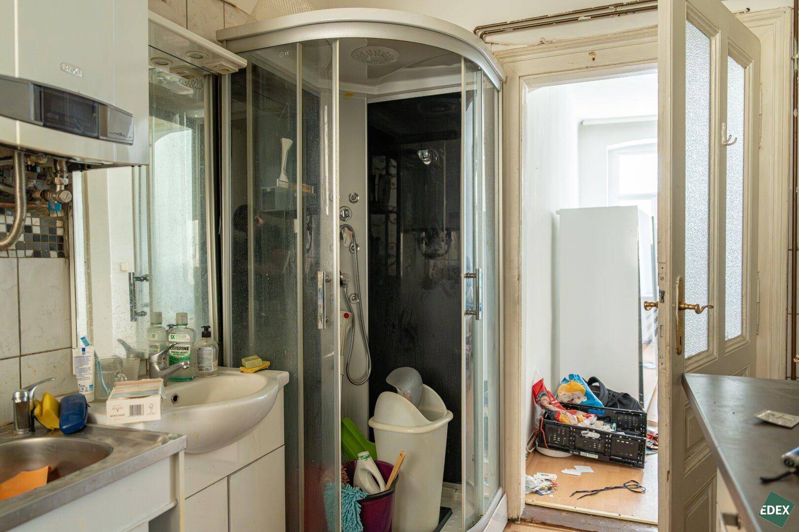 Badezimmer mit Eckduschkabine, Waschbecken und Warmwasserbereiter, in renovierungsbedürftigem Zustand.