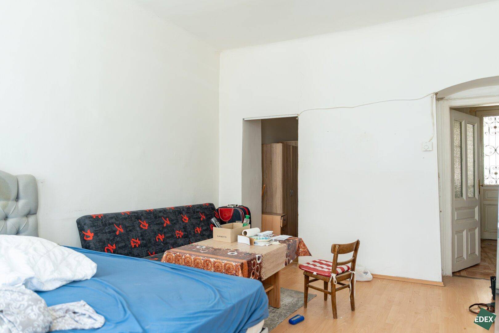 Wohnbereich mit Sofa, Bett und Tisch, der durch eine offene Tür mit einem weiteren Raum verbunden ist.