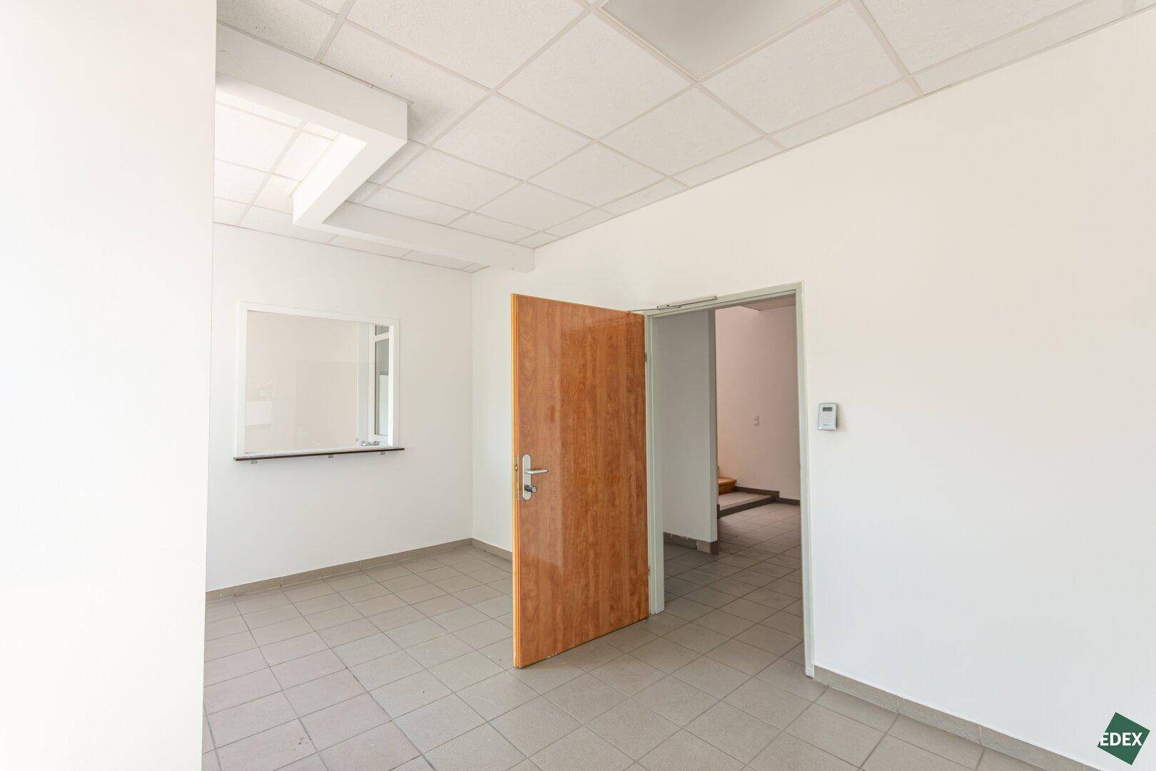 Büroraum mit Fliesenboden und Holztür, die zu einem weiteren Bereich führt, mit kleiner Durchreiche.