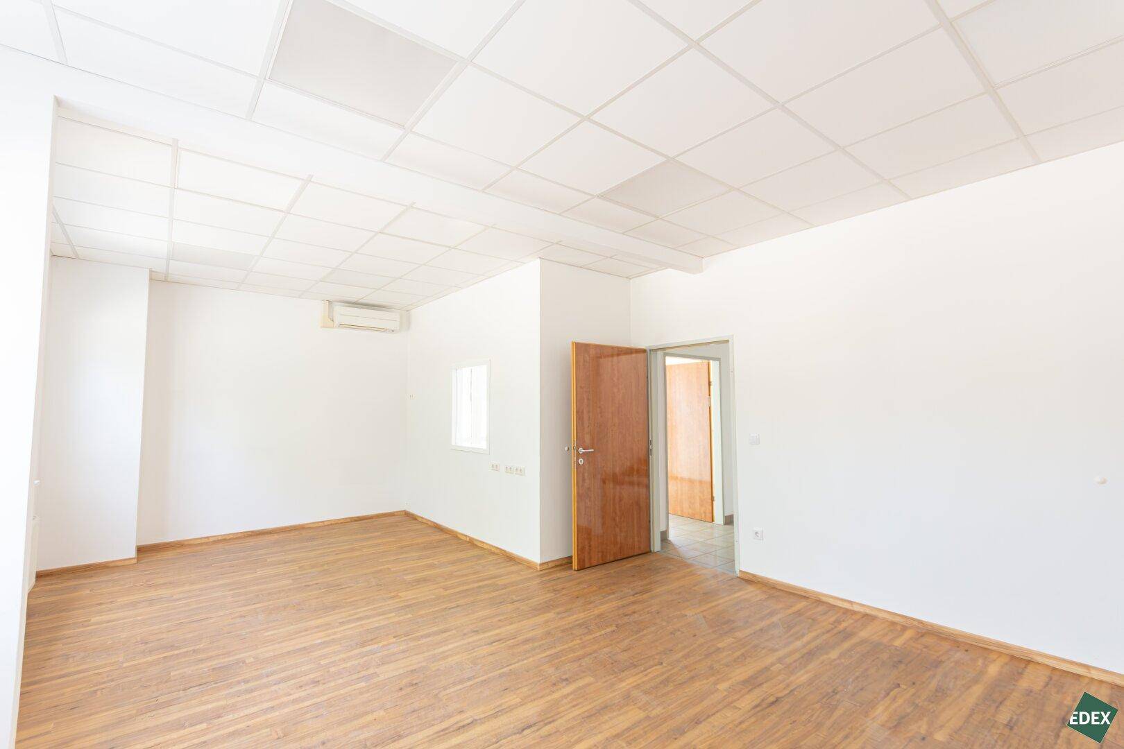 Heller Büroraum mit Laminatboden und Klimaanlage, der durch eine Holztür zugänglich ist.