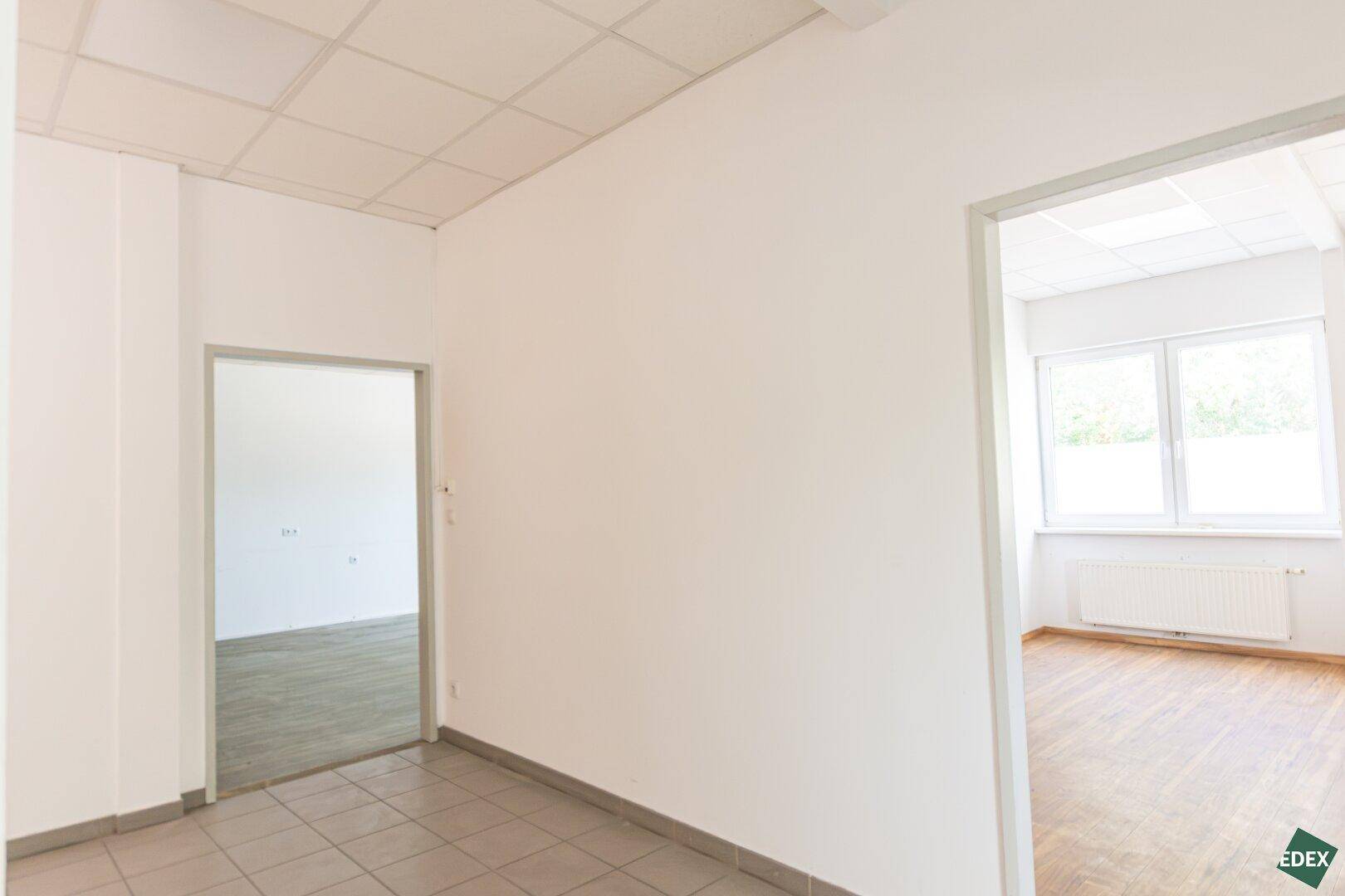 Flur mit Fliesenboden, weißen Wänden und Blick in angrenzende Büroräume.