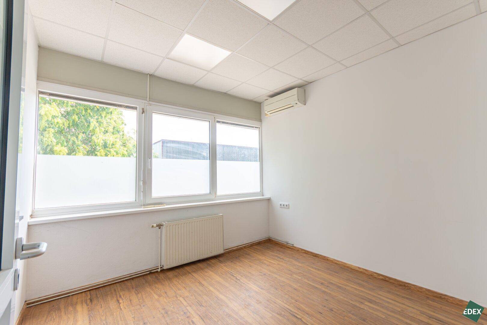 Heller Büroraum mit großen Fenstern, Laminatboden und Klimaanlage, bietet viel Tageslicht.