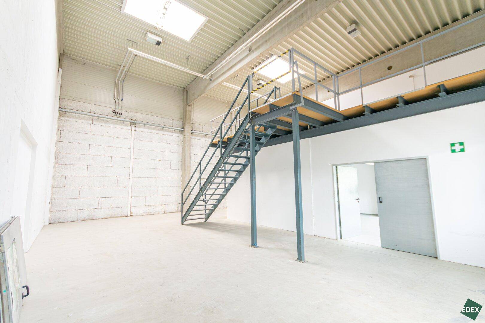 Hohe Lagerhalle mit Betonwänden, Metalltreppe zu einer Empore und Oberlichtern.