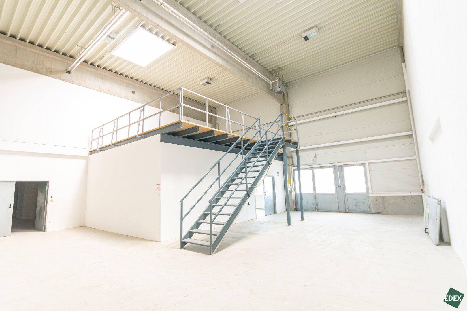 Weitläufige Lagerhalle mit hoher Decke, Metalltreppe zur Empore und großen Toren.
