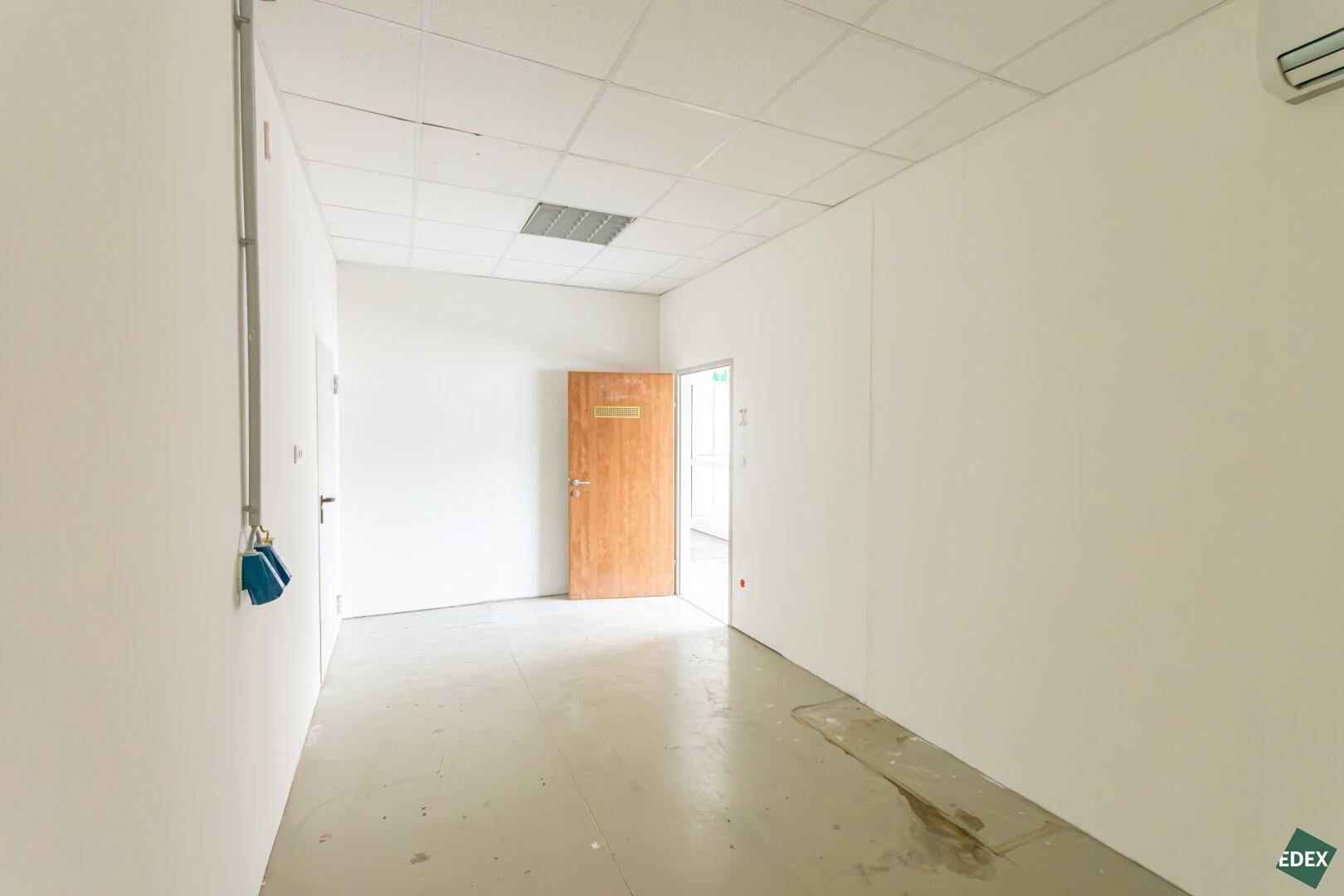 Schmaler Flur mit Betonboden, weißen Wänden und Holztür zu einem weiteren Raum.