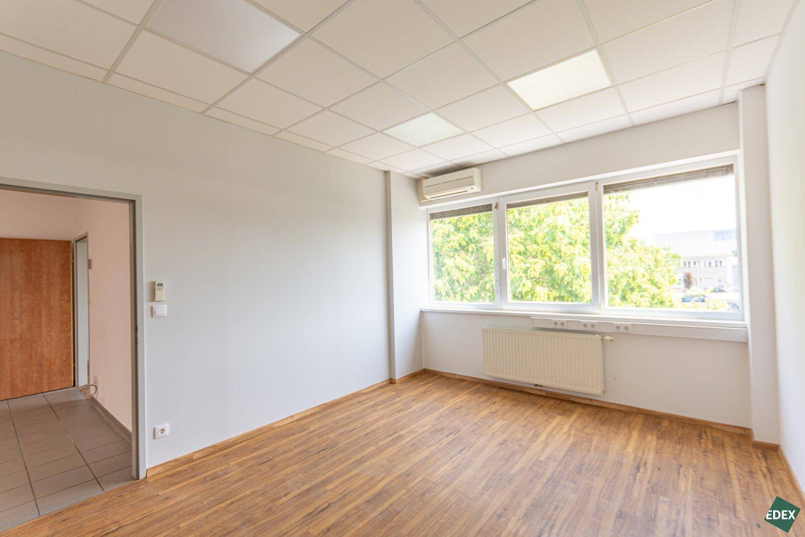 Büroraum mit Laminatboden und großen Fenstern, die einen Blick ins Grüne ermöglichen.
