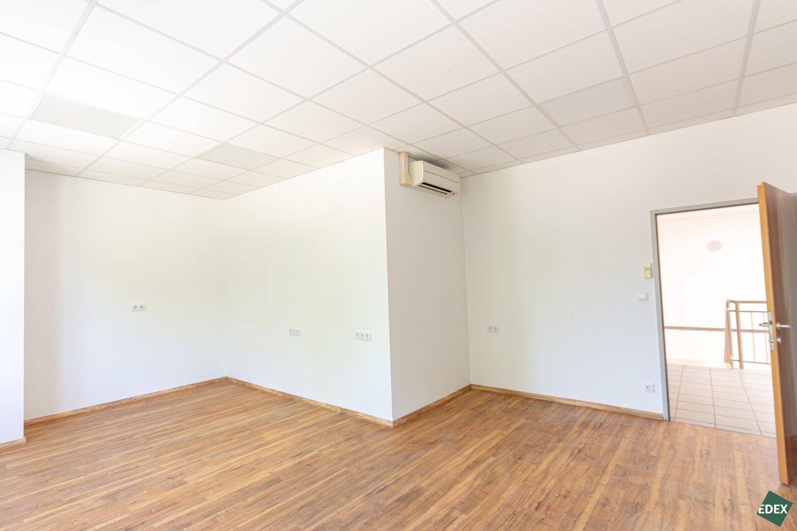 Büroraum mit Laminatboden und Klimaanlage, der durch eine offene Tür zugänglich ist.