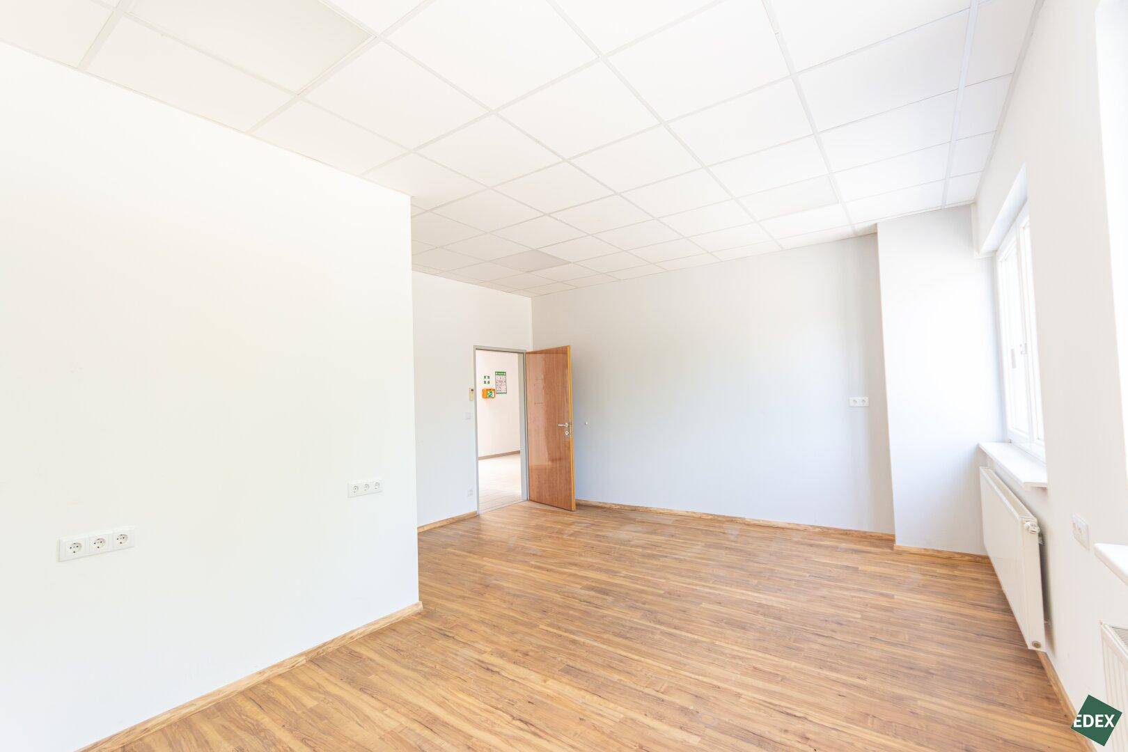 Leerer Büroraum mit Laminatboden und Fenster, das viel Tageslicht hereinlässt.