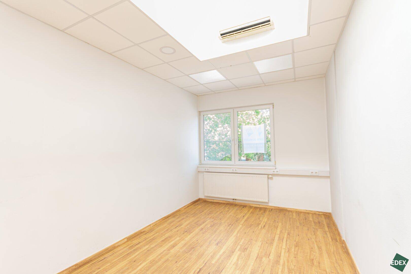 Kleinerer Büroraum mit Fenster, Laminatboden und Klimaanlage, ideal für Einzelbüros.