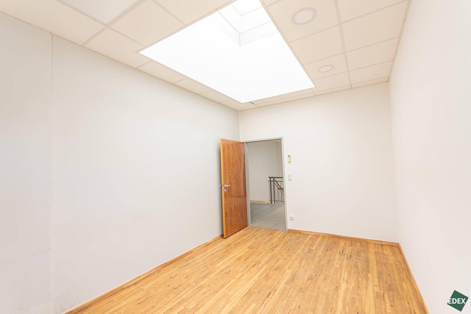 Büroraum mit Laminatboden, Holztür und Deckenfenster, das für zusätzliches Tageslicht sorgt.