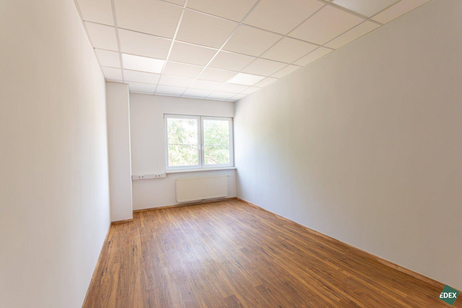 Heller Büroraum mit großem Fenster, Laminatboden und Heizkörper, ideal für konzentriertes Arbeiten.