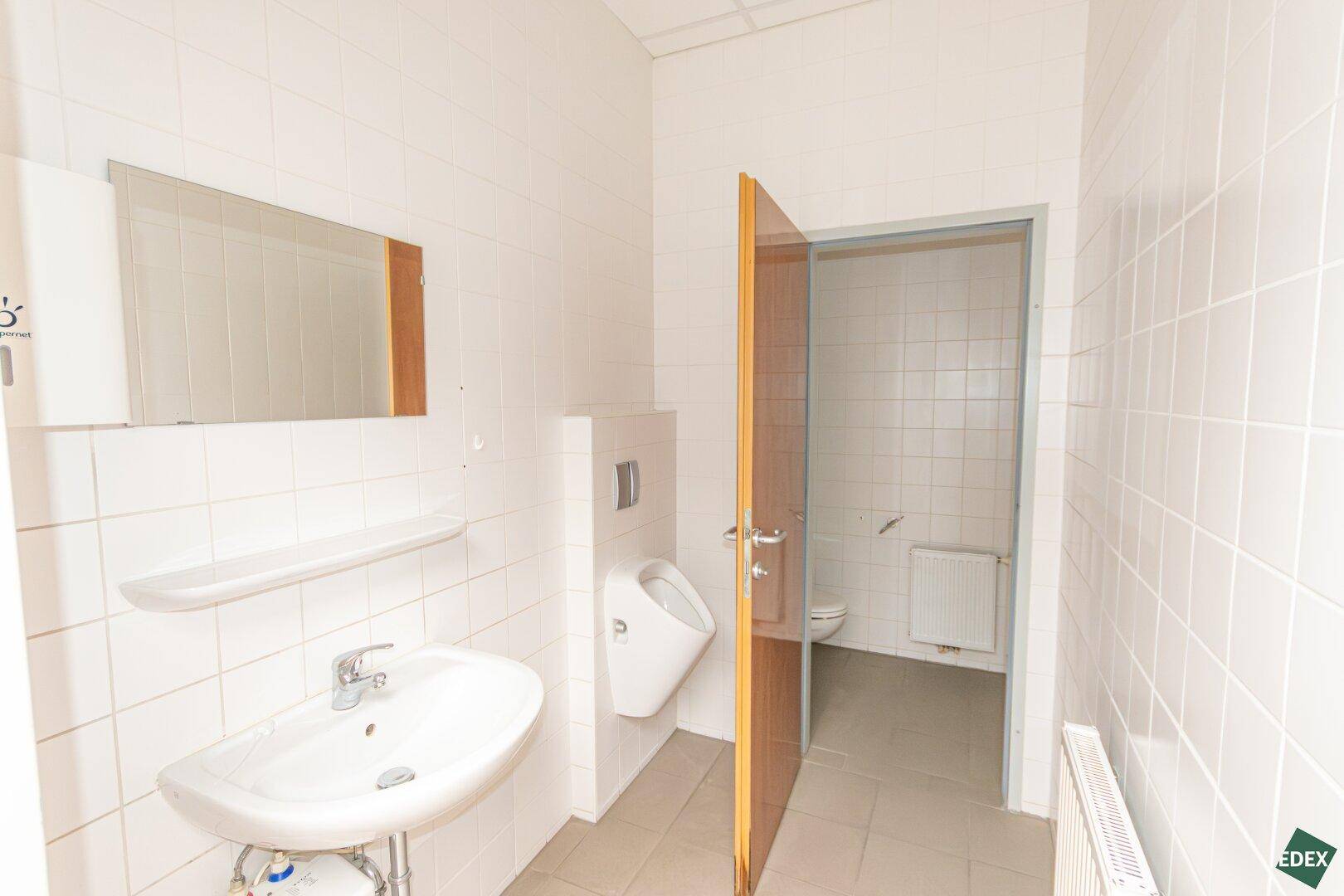 Helles Badezimmer mit weißen Fliesen, Waschbecken, Urinal und Toilette im Nebenraum.