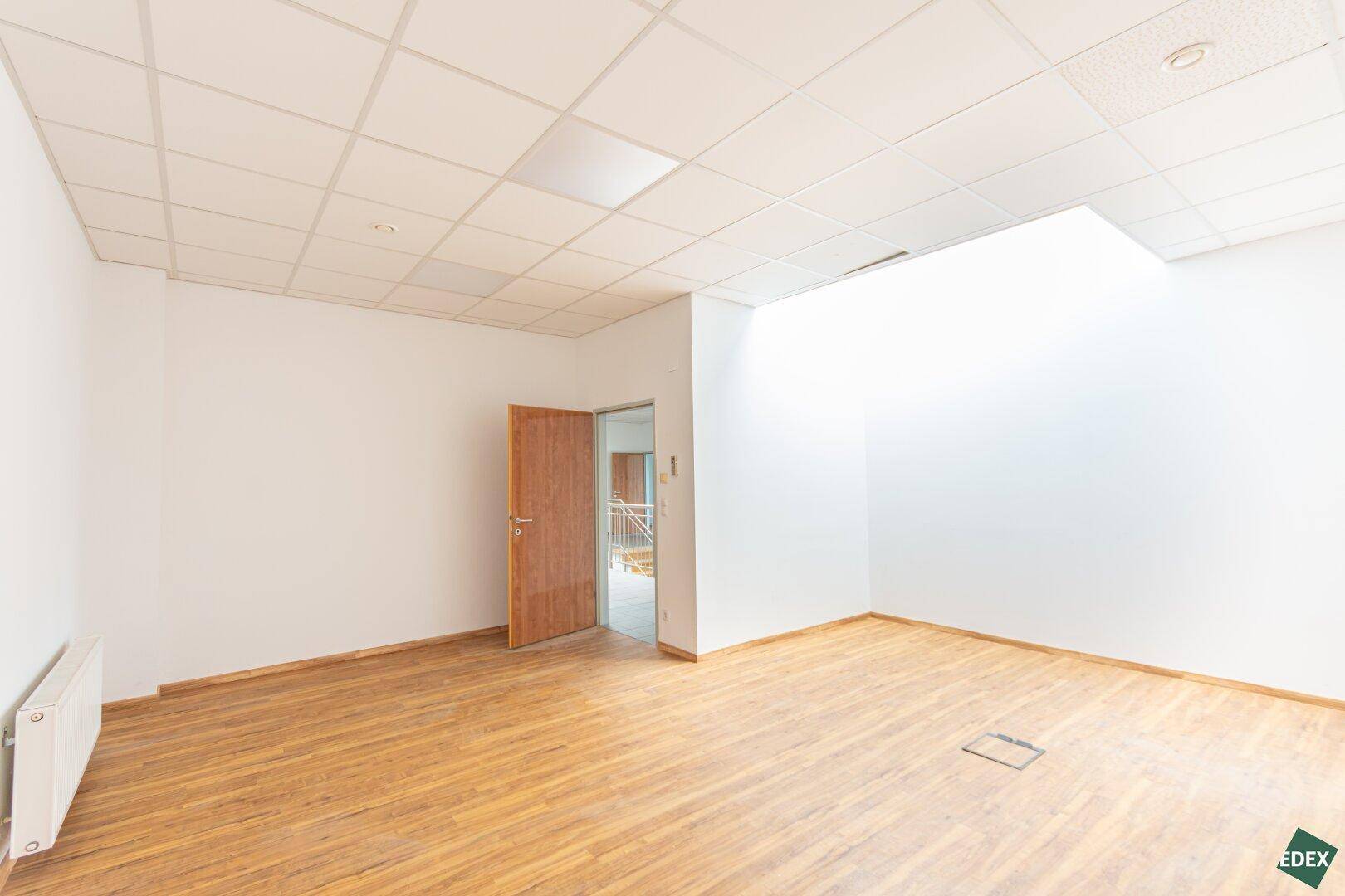 Leerer Büroraum mit Laminatboden und Holztür, der Zugang zu weiteren Bereichen bietet.