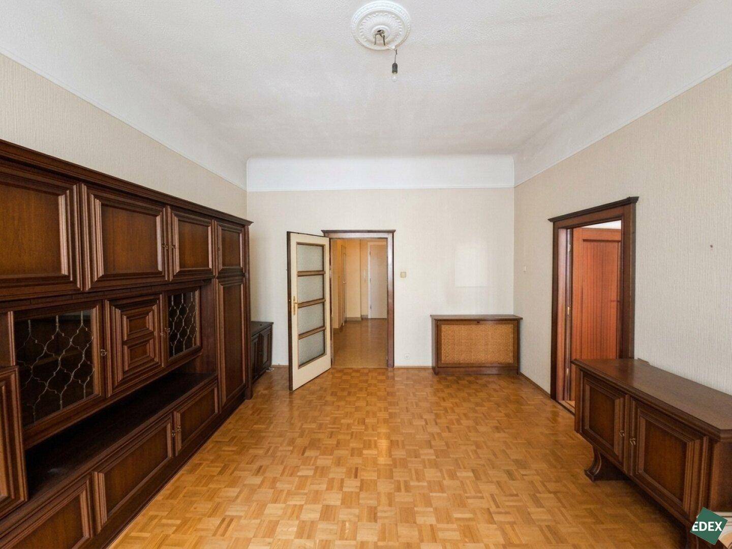 Wohnzimmer mit dunklen Holzmöbeln, Parkettboden und Zugang zu weiteren Räumen.