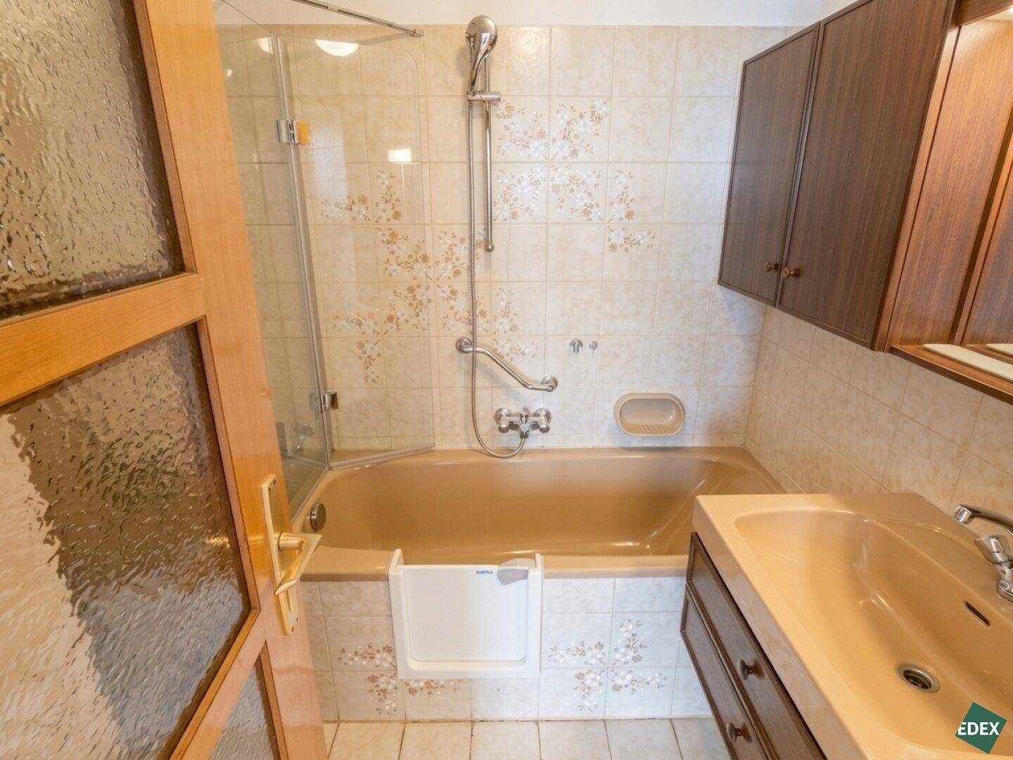 Badezimmer im 80er-Jahre-Stil mit beiger Badewanne, Duschvorrichtung und gemusterten Fliesen.