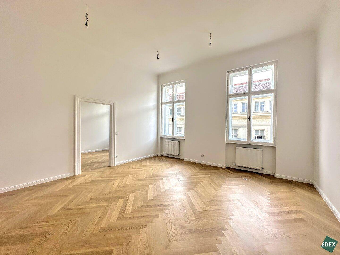 Geräumiger, leerer Raum mit Fischgrätparkett und zwei hohen Fenstern, bietet viel Tageslicht.