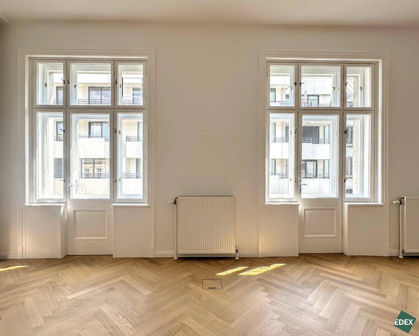 Zwei hohe Fenster in einem hellen Raum mit Fischgrätparkett, die viel Sonnenlicht hereinlassen.