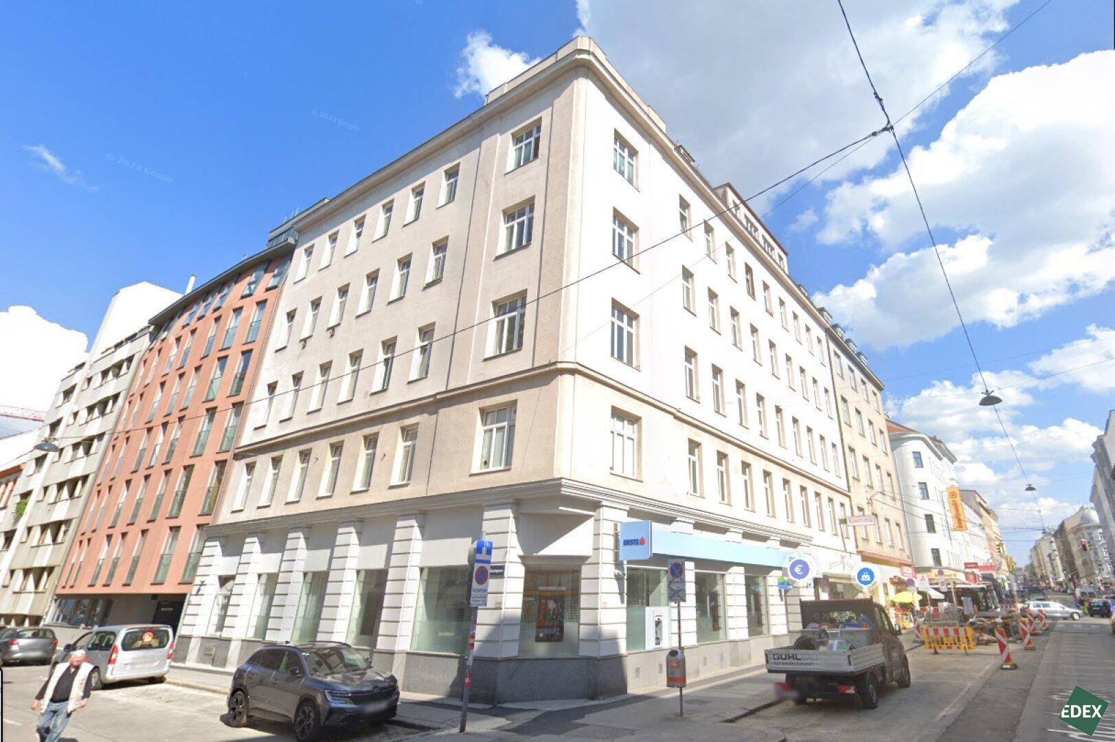 Eckgebäude in einer städtischen Umgebung mit vielen Fenstern und Geschäften im Erdgeschoss.