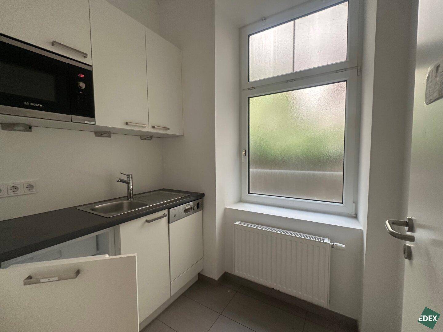 Kompakte Küchenzeile mit weißen Schränken, integrierter Mikrowelle und Spülmaschine sowie einem Fenster.
