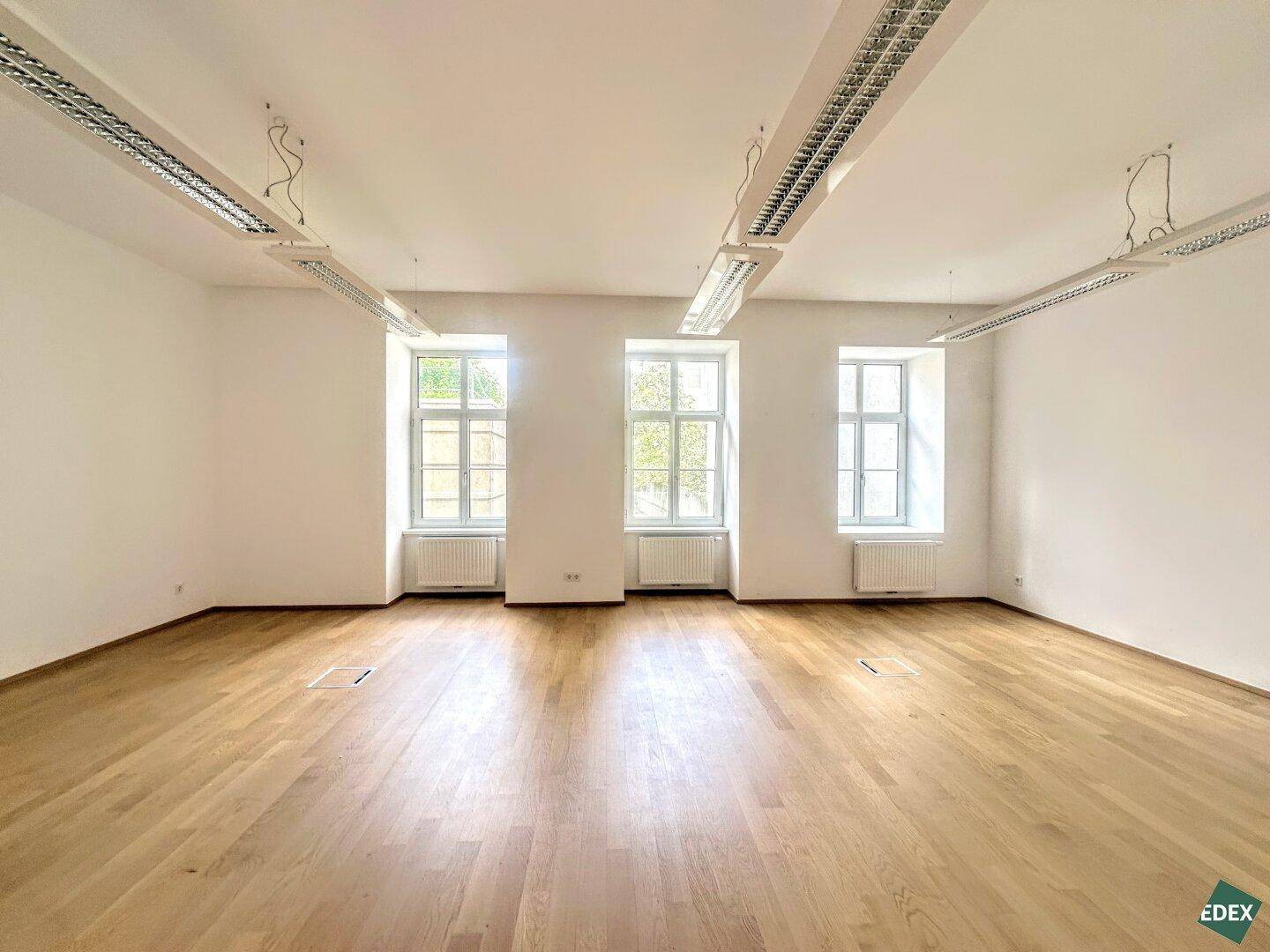 Heller und leerer Büroraum mit drei großen Fenstern, Holzboden und Deckenleuchten, ideal für flexible Gestaltung.