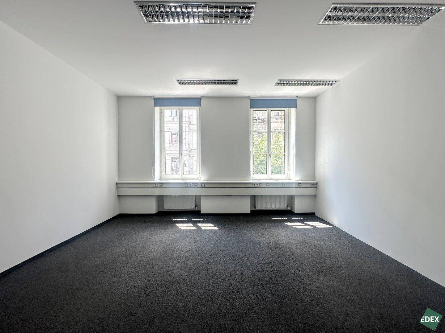 Helles Einzelbüro mit Teppichboden, zwei großen Fenstern und blauen Rollos, bietet viel Tageslicht.