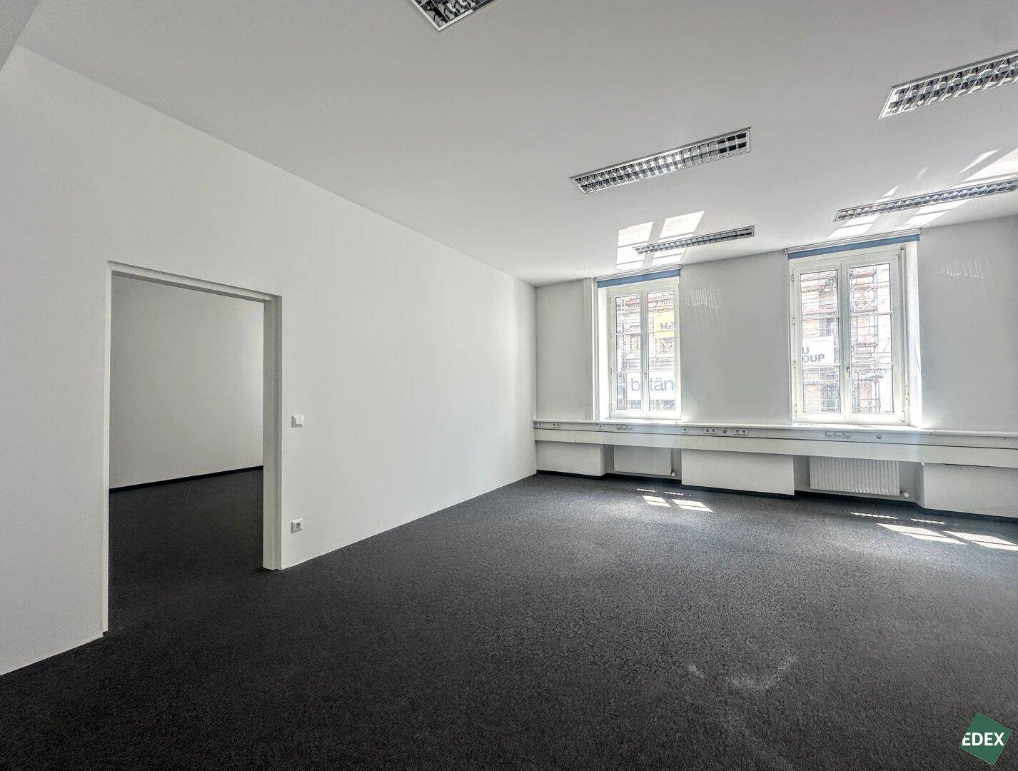 Bürofläche mit Teppichboden und Zugang zu einem weiteren Raum, beleuchtet durch große Fenster.