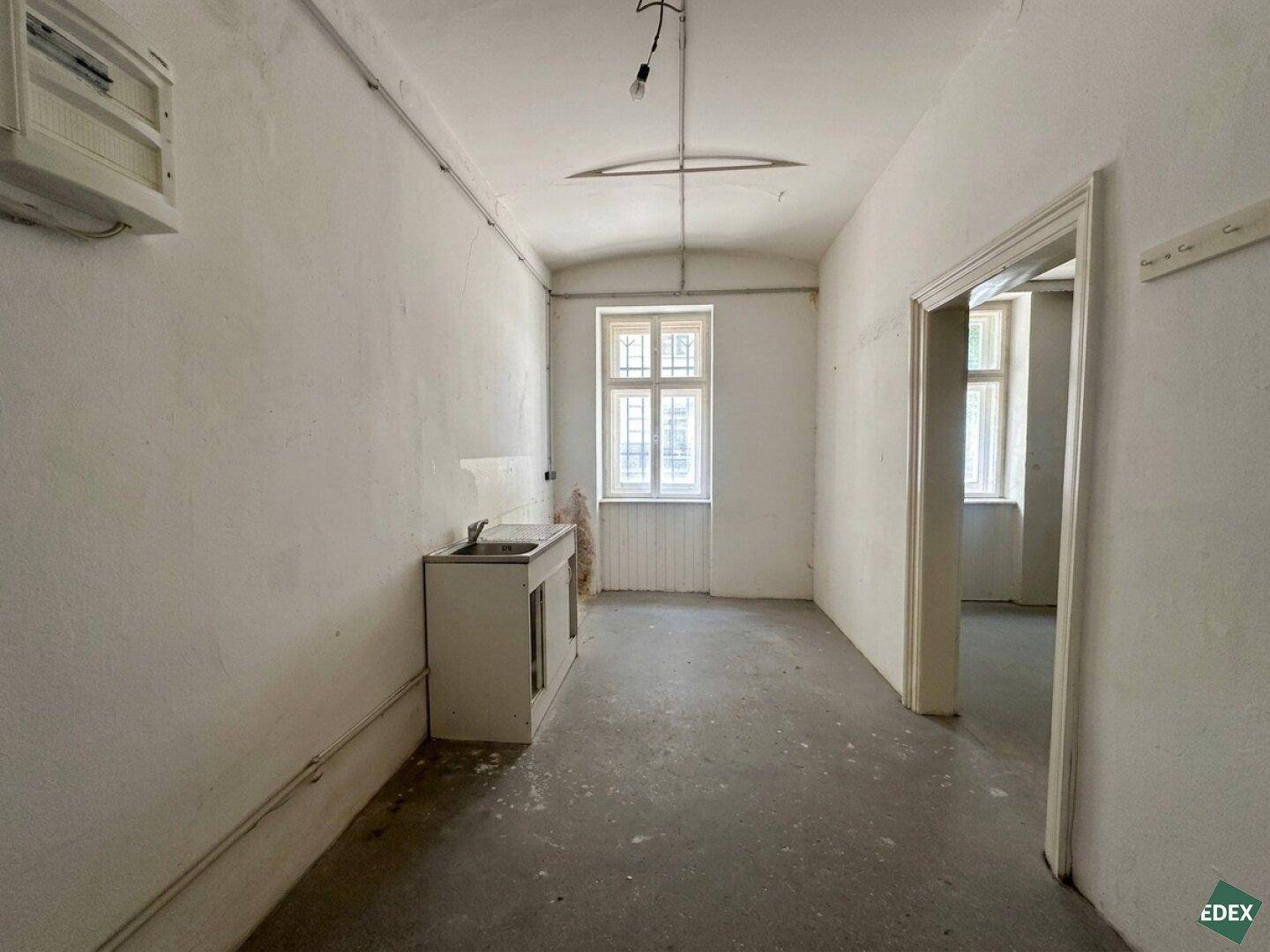 Langer Raum mit einem einfachen Waschbecken, Betonboden und einem Fenster, das Licht spendet.