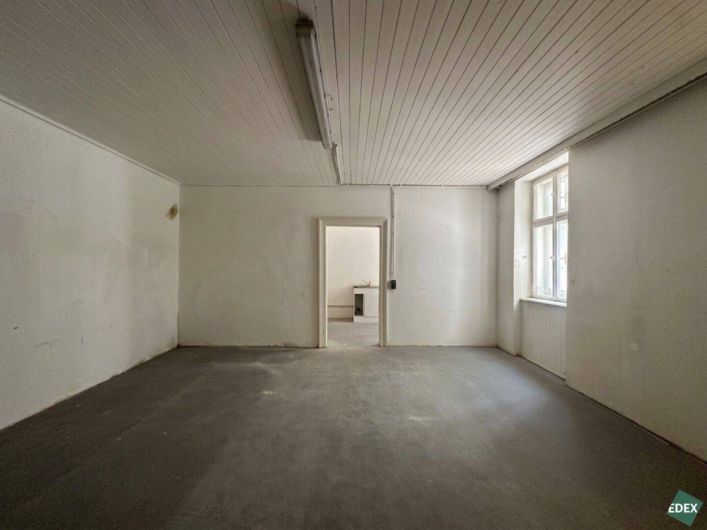 Heller, leerer Raum mit Betonboden, weißer Decke und einem Fenster, das Tageslicht hereinlässt.