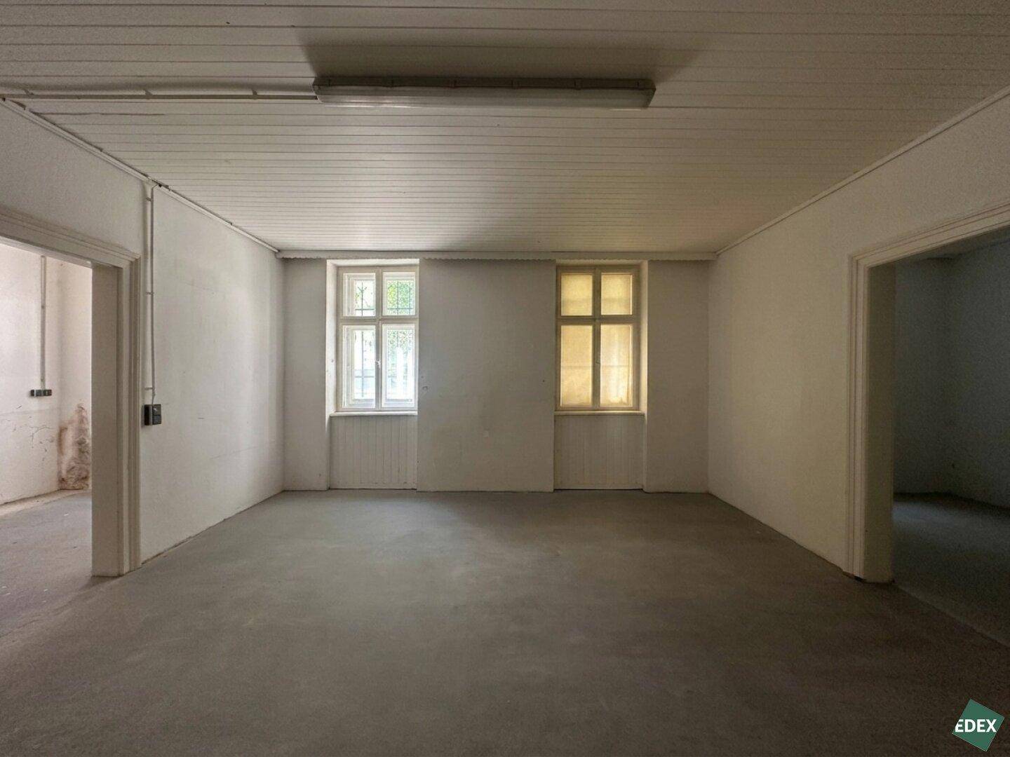 Geräumiger, leerer Raum mit zwei großen Fenstern, Betonboden und einer Holzpaneeldecke.