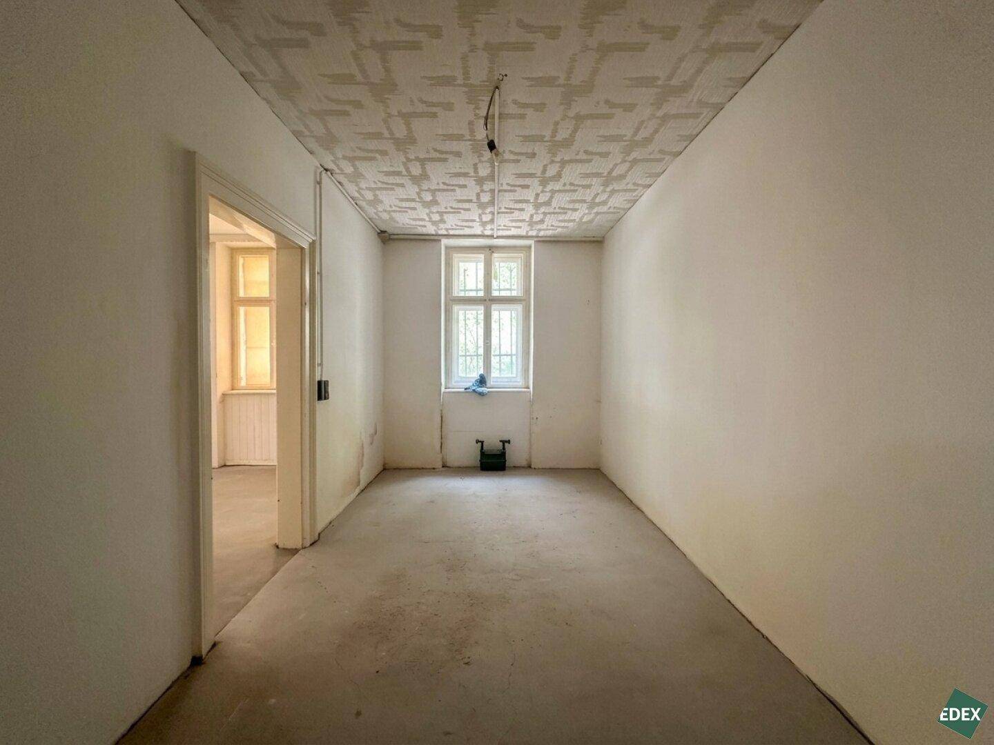 Leerer Raum mit Betonboden, einem Fenster und einer strukturierten Decke, die renoviert werden muss.