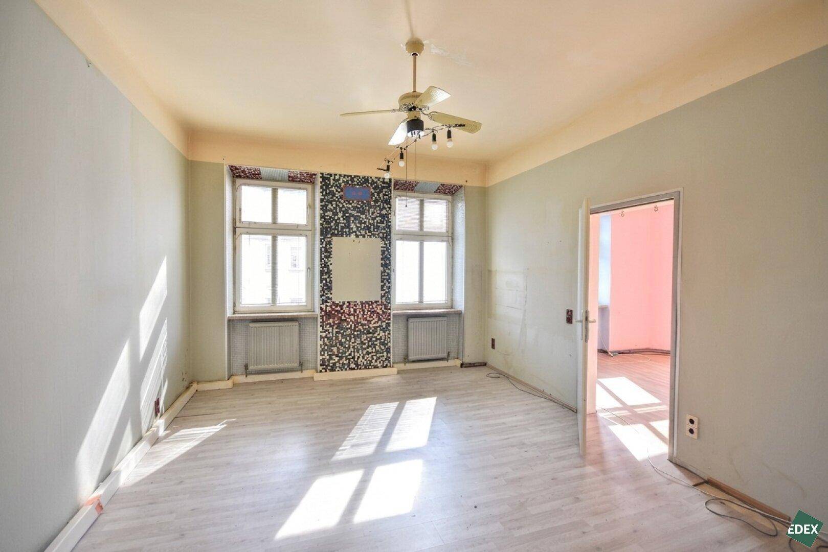 Geräumiges, unmöbliertes Zimmer mit großen Fenstern, Deckenventilator und Renovierungsbedarf.
