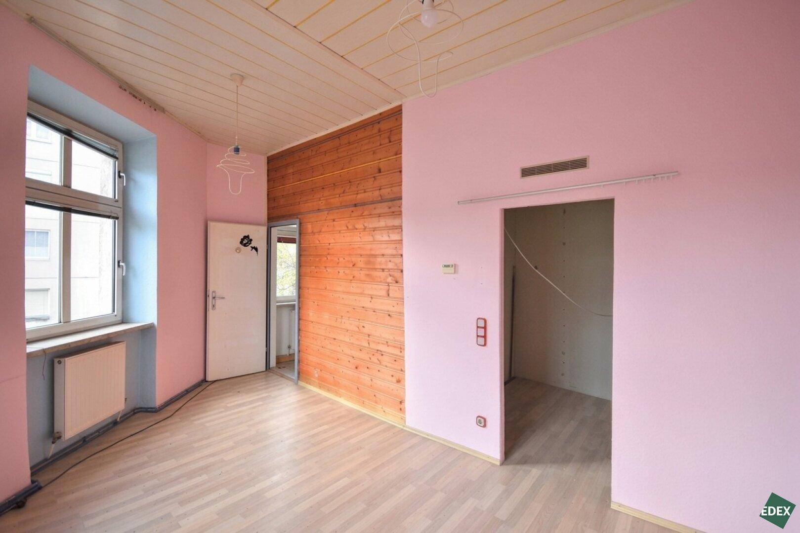 Unmöbliertes Zimmer mit rosa Wänden, Holzverkleidung, Laminatboden und Blick ins Grüne.