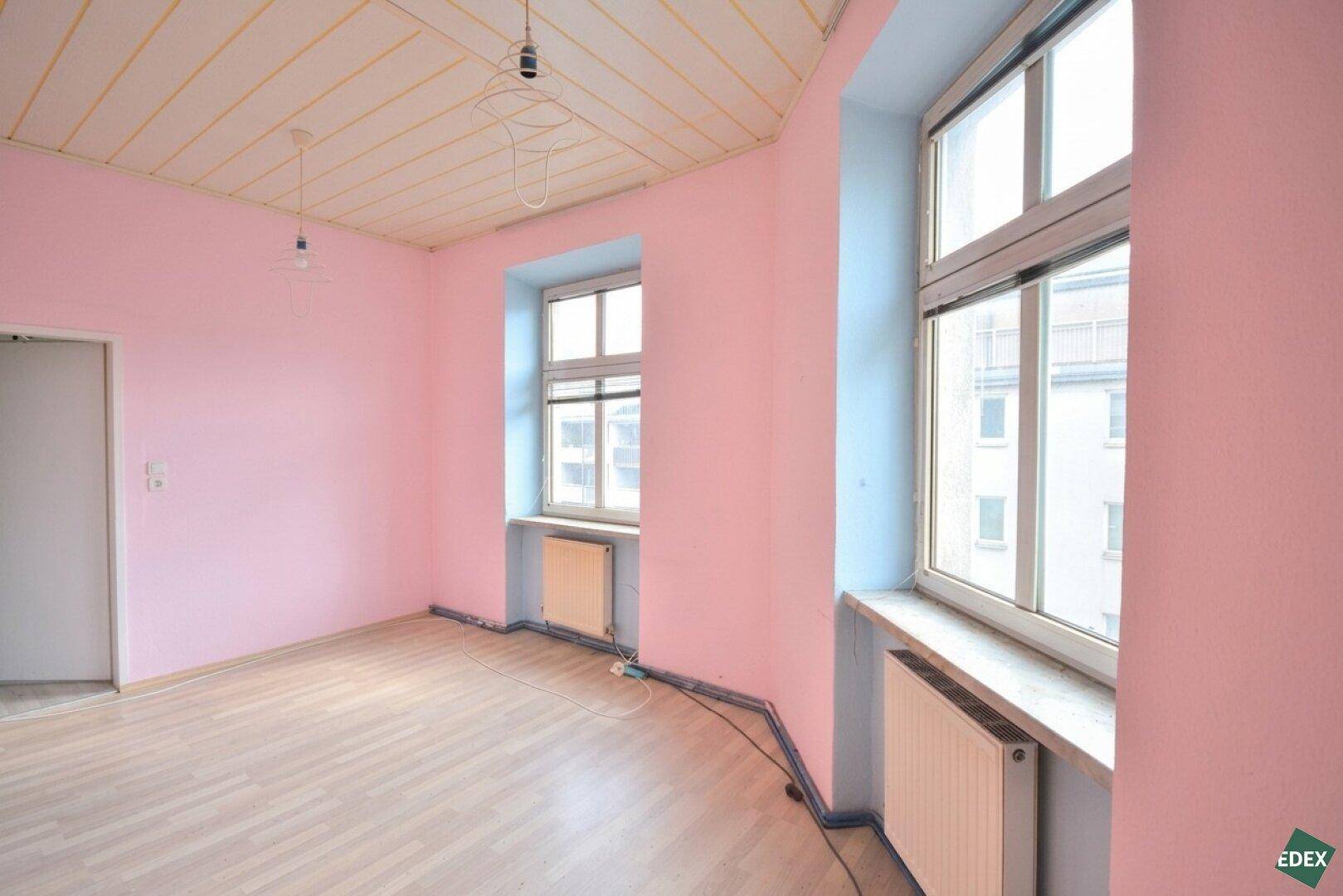Heller Raum mit rosa Wänden, Laminatboden, zwei Fenstern und Blick auf die Stadt.