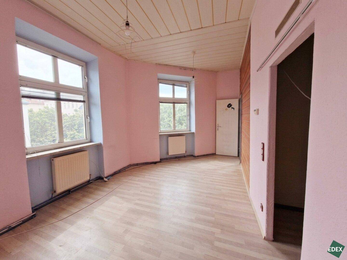 Leeres Zimmer mit rosa Wänden, Laminatboden, zwei Fenstern und Blick ins Grüne.