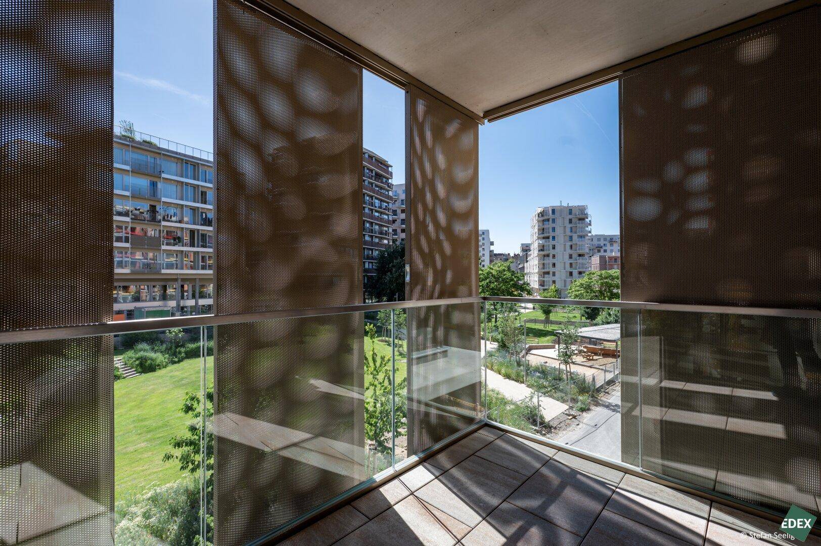 Moderner Balkon mit Fliesenboden, Glasgeländer und perforierten Sichtschutzpaneelen mit Grünblick.