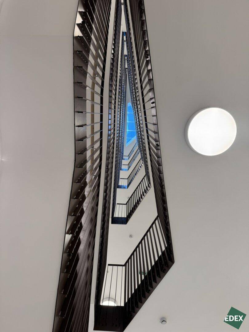 Blick nach oben in ein modernes Treppenhaus mit geometrischen Metallgeländern und einem hellen Oberlicht.