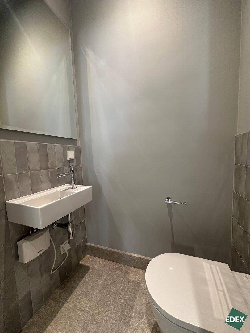 Modernes Gäste-WC mit Wand-WC, kleinem Waschbecken und grauen Fliesen an Wänden und Boden.